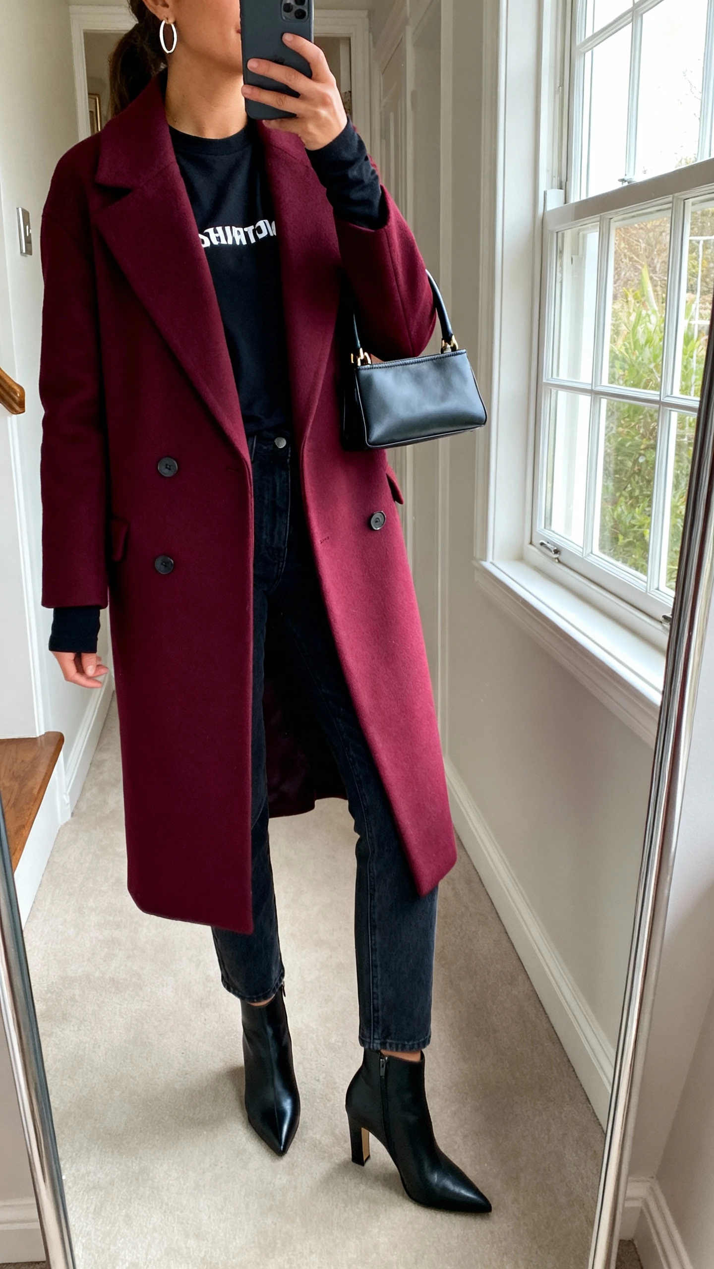 iPhone mirror selfie of a woman wearing a bold statement coat in deep burgundy over a simple base of a black long-sleeve tee and dark straight-leg jeans, sleek heeled booties, minimal hoop earrings, small top-handle bag, face not visible, side angle, entryway full-length mirror with natural window light, iPhone photo quality.