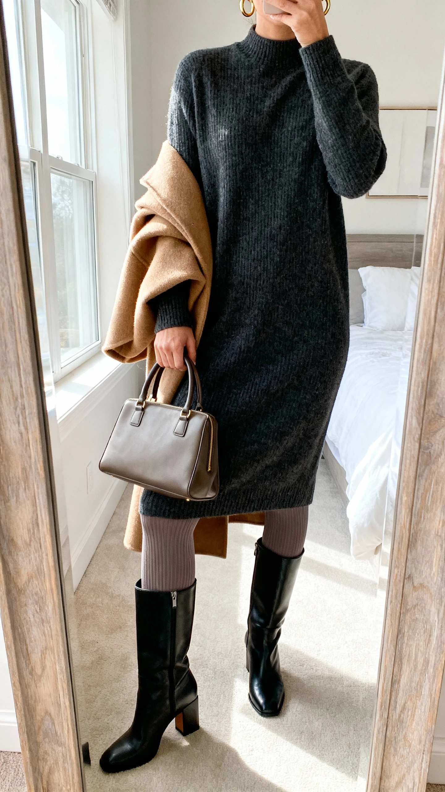iPhone mirror selfie of a woman wearing a charcoal midi sweater dress, opaque thermal tights, knee-high black leather boots with a block heel, a camel wool coat draped over her shoulders, minimal gold hoops, and a structured top-handle tote; face not visible, cropped at neck, bedroom mirror, natural window light, iPhone photo quality.