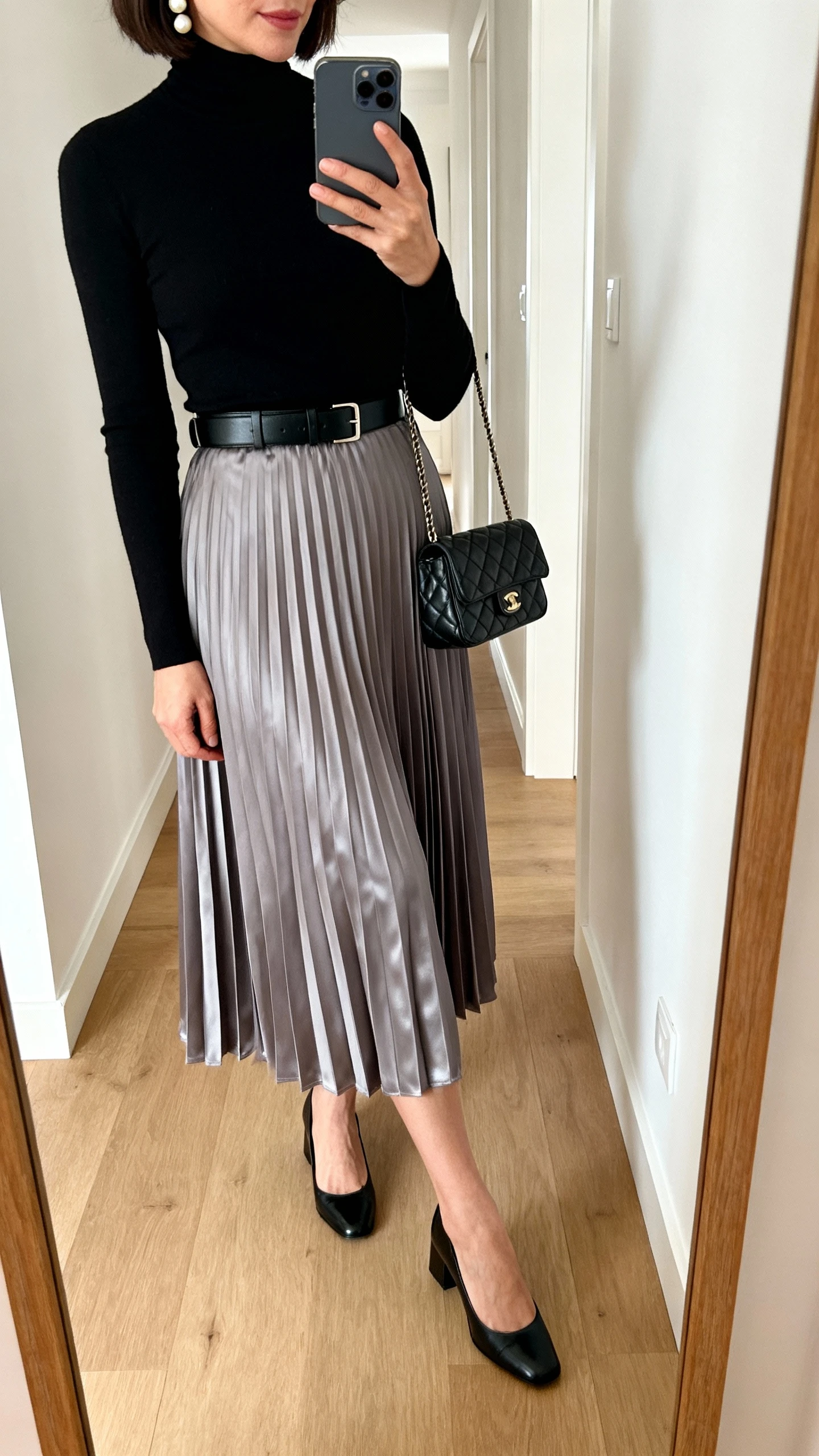 iPhone side-angle mirror selfie of a woman wearing a pleated midi skirt in satin dove gray with a fitted black turtleneck, classic black belt, low block-heel pumps, pearl studs, small quilted shoulder bag, face not visible, cropped at shoulders, hallway mirror with natural daylight, iPhone photo quality.