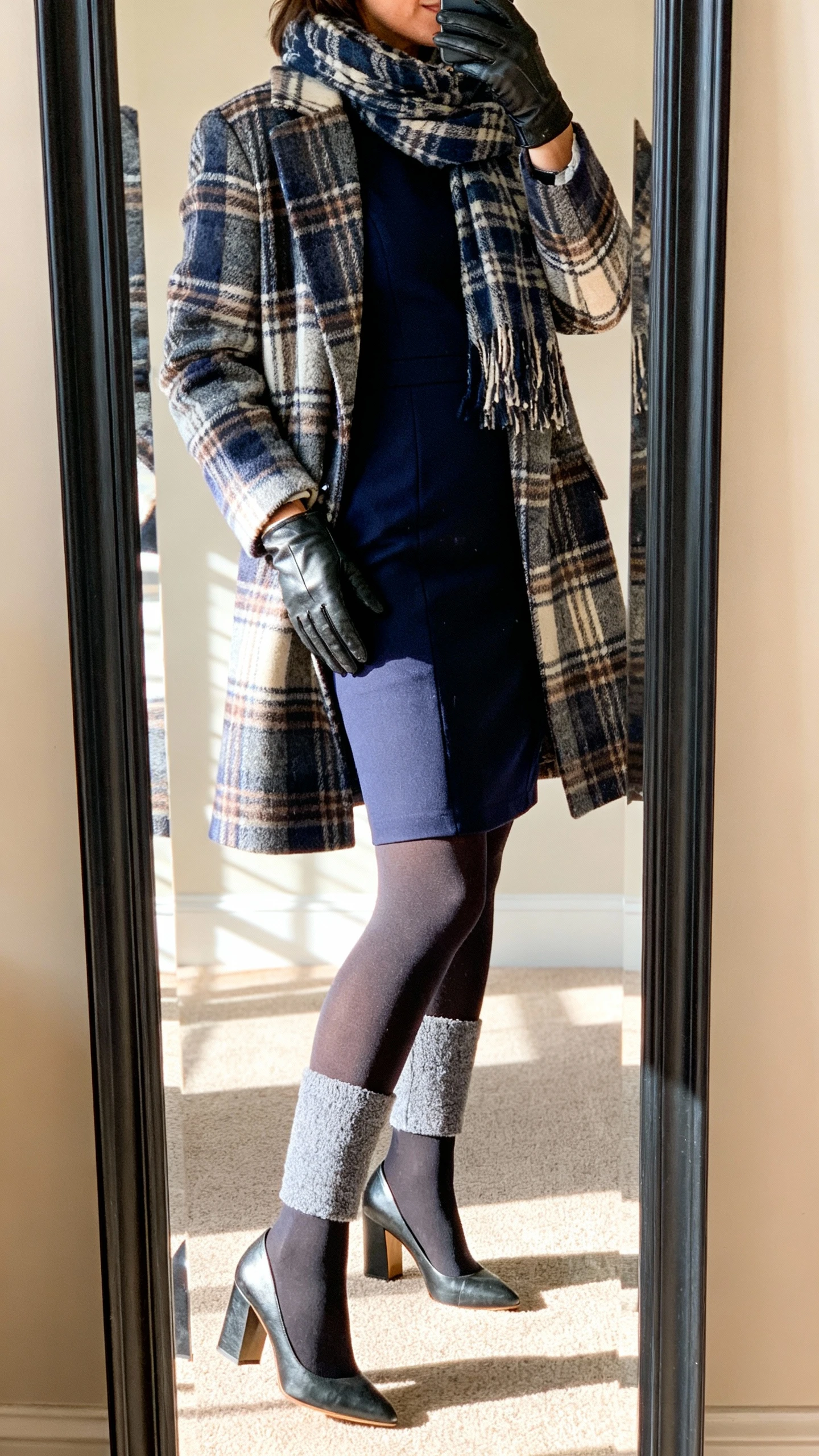 iPhone side-angle mirror selfie of a woman wearing a navy structured sheath dress, opaque fleece-lined tights, block-heel pumps, a plaid statement coat, leather gloves, and a matching scarf; face not visible, full-length mirror, indoor afternoon light, iPhone photo quality.
